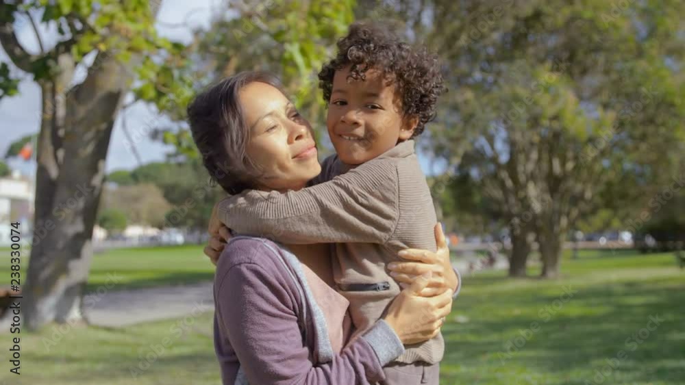 Curly African American son hugging and kissing mother in slow motion. Cheerful Asian mom embracing little girl in summer park. Parenthood concept Curly African American son hugging and kissing mother in slow motion. Cheerful Asian mom embracing little girl in summer park. Parenthood concept