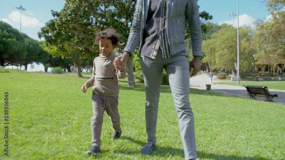 African American little boy holding hands with father and walking in park. Front view of dad and son walking on green grass in slow motion. Happy family concept