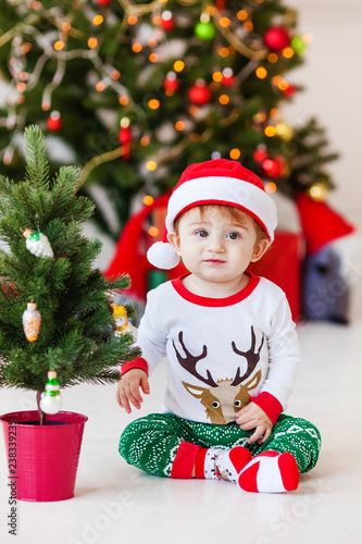 Cute little santa baby in white and green pajamas. Christmas tree and new year gifts on the background