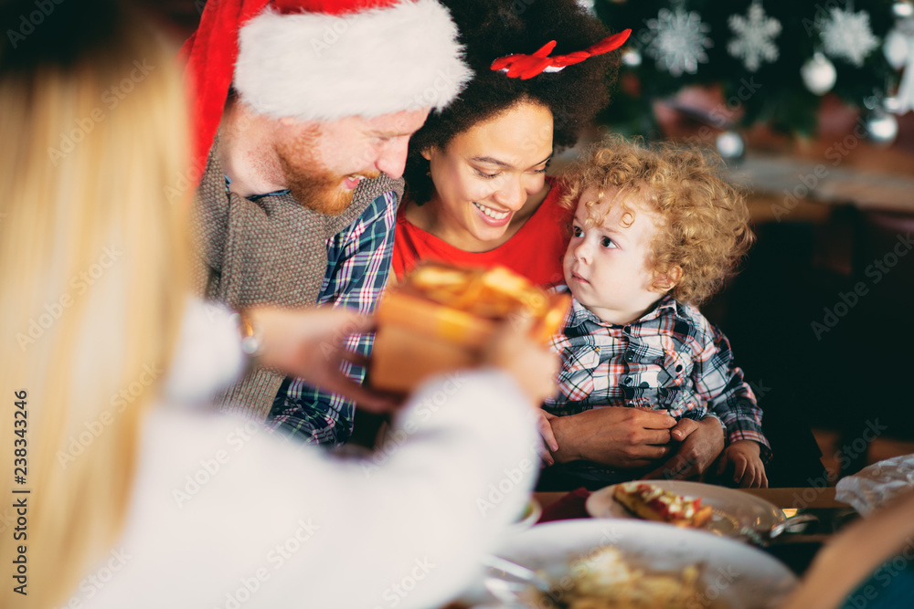 Man and mixed race woman giving toddler Christmas gift while sitting at table. Christmas holidays concept. Man and mixed race woman giving toddler Christmas gift while sitting at table. Christmas holidays concept.