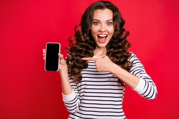 Portrait of nice cute crazy funny attractive amazed wondered stunned wavy-haired lady wearing striped pullover pointing at black screen display isolated over bright vivid shine red background