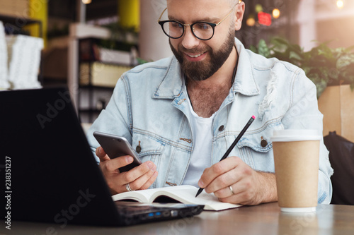 Young bearded man trendy glasses sits cafe in front of laptop computer, uses smartphone, takes notes in notebook.