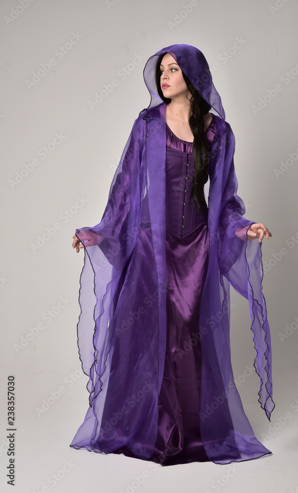 full length portrait of beautiful girl with long black hair, wearing purple fantasy medieval gown and cloak. standing pose on grey studio background. full length portrait of beautiful girl with long black hair, wearing purple fantasy medieval gown and cloak. standing pose on grey studio background.