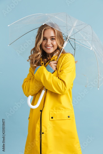 Full length image of charming woman 20s wearing yellow raincoat standing under transparent umbrella, isolated over blue background