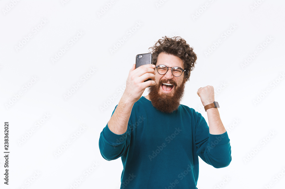 Naklejka premium Amazed and cheerful bearded man looking at smartphone and celebrating success over white background