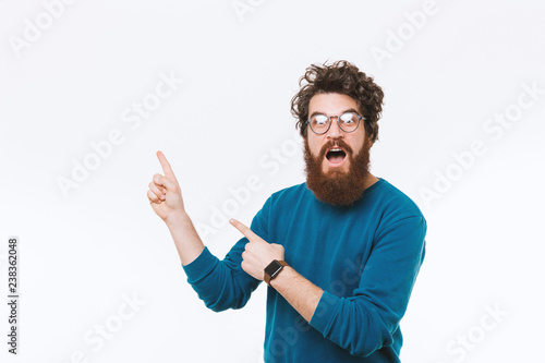 Amazed bearded man wearing glasses and pointing away over white background photo