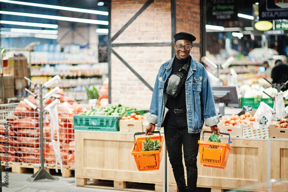 Stylish casual african american man at jeans jacket and black beret holding two baskets, walking and shopping at supermarket.