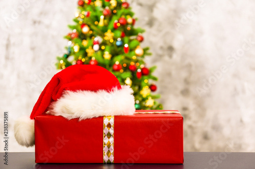 Festive composition with one single present in colorful wrapping paper with santa claus hat on foreground and blurry decorated Christmas spruce tree on background. Close up, copy space.