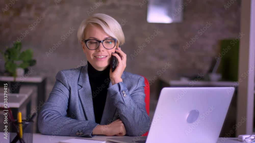 Portrait of middle-aged blonde short-haired businesswoman in glasses gladly talking on cellphone in office. Portrait of middle-aged blonde short-haired businesswoman in glasses gladly talking on cellphone in office.