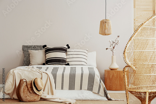 Copy space on white empty wall of bright trendy bedroom with flower in vase on wooden nightstand, king size bed with striped bedding and rattan peacock chair photo