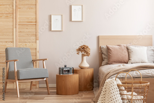 Trendy retro armchair next to two round wooden tables with vintage camera and vase with flowers in beige bedroom interior