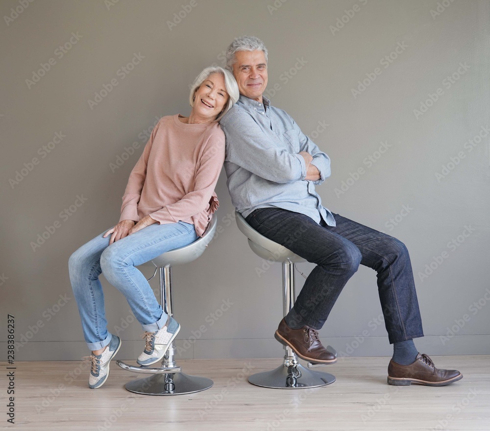  Happy senior couple sitting on bar stools on grey backgound - obrazy, fototapety, plakaty 