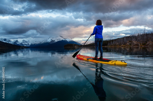 Girl Paddle Boarding in a peaceful and calm glacier lake during a vibrant cloudy sunset. Taken in Maligne Lake, Jasper National Park, Alberta, Canada.