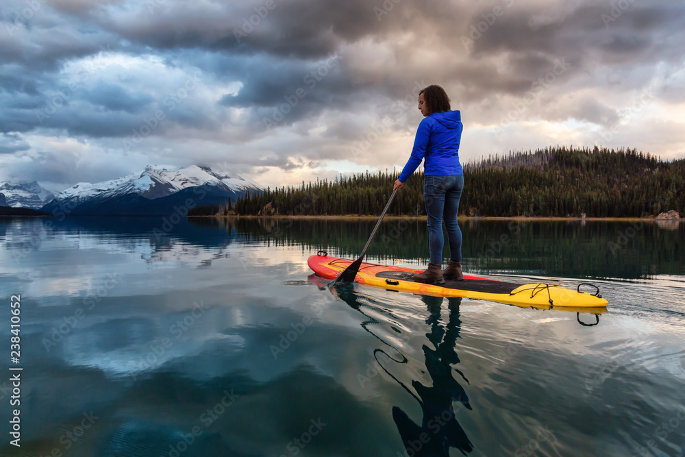 Girl Paddle Boarding in a peaceful and calm glacier lake during a vibrant cloudy sunset. Taken in Maligne Lake, Jasper National Park, Alberta, Canada. Girl Paddle Boarding in a peaceful and calm glacier lake during a vibrant cloudy sunset. Taken in Maligne Lake, Jasper National Park, Alberta, Canada.