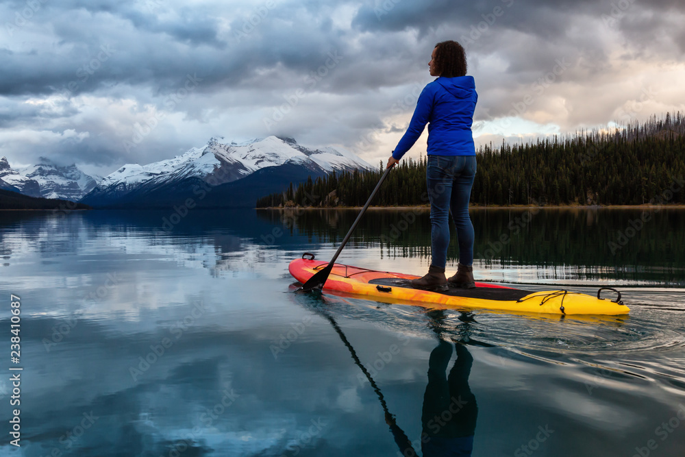 Girl Paddle Boarding in a peaceful and calm glacier lake during a vibrant cloudy sunset. Taken in Maligne Lake, Jasper National Park, Alberta, Canada. Girl Paddle Boarding in a peaceful and calm glacier lake during a vibrant cloudy sunset. Taken in Maligne Lake, Jasper National Park, Alberta, Canada.