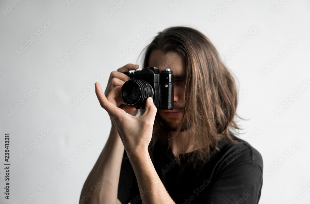 Handsome young bearded man with a long hair and in a black shirt holding vintage old-fashioned film camera on a white background and looking in camera viewfinder. Handsome young bearded man with a long hair and in a black shirt holding vintage old-fashioned film camera on a white background and looking in camera viewfinder.