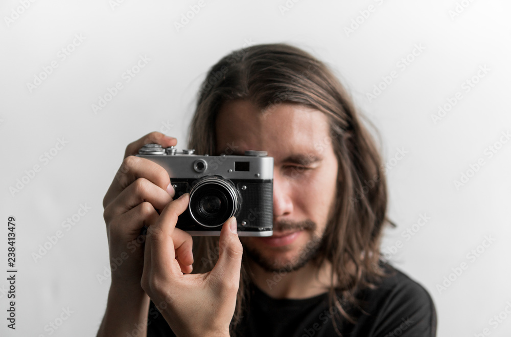 Handsome young bearded man with a long hair and in a black shirt holding vintage old-fashioned film camera on a white background and looking in camera viewfinder. Handsome young bearded man with a long hair and in a black shirt holding vintage old-fashioned film camera on a white background and looking in camera viewfinder.