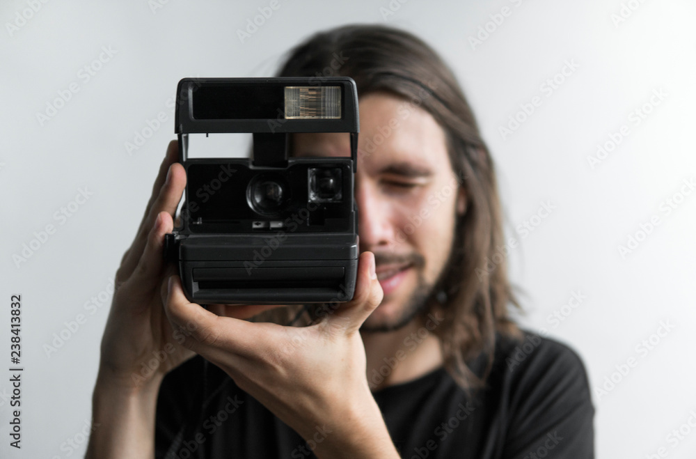 Handsome young bearded man with a long hair and in a black shirt holding vintage old-fashioned film camera on a white background and looking in camera viewfinder.