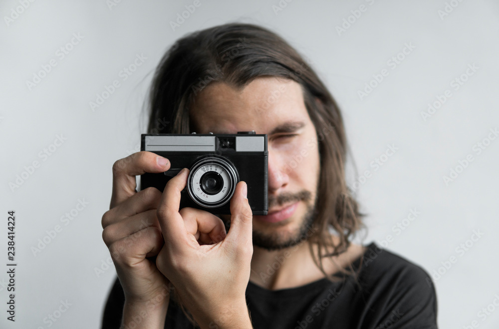 Handsome young bearded man with a long hair and in a black shirt holding vintage old-fashioned film camera on a white background and looking in camera viewfinder. Handsome young bearded man with a long hair and in a black shirt holding vintage old-fashioned film camera on a white background and looking in camera viewfinder.