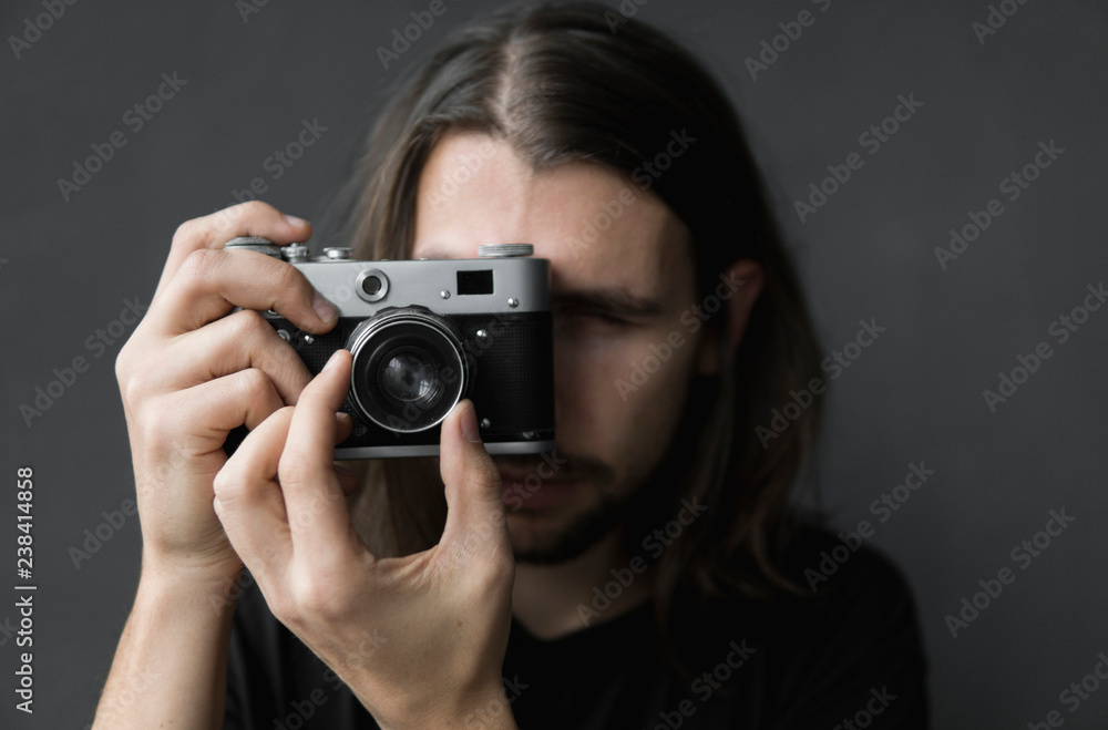 Handsome young bearded man with a long hair and in a black shirt holding vintage old-fashioned film camera on a black background and looking in camera viewfinder. Handsome young bearded man with a long hair and in a black shirt holding vintage old-fashioned film camera on a black background and looking in camera viewfinder.