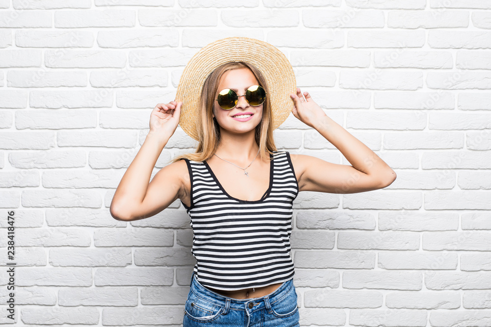 Young happy woman smiling in sunglasses and summer hat over white brick wall