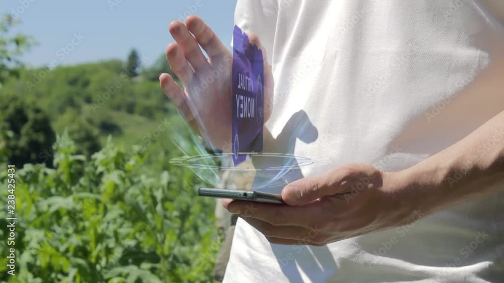 Man shows concept hologram Virtual money on his phone. Person in white t-shirt with future technology holographic screen and green nature background Man shows concept hologram Virtual money on his phone. Person in white t-shirt with future technology holographic screen and green nature background