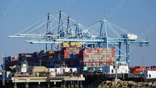 Container ship in a modern industrial shipping harbor operation in the Port of Los Angeles by a row of gantry cranes, surrounded by other port and ocean vessels.