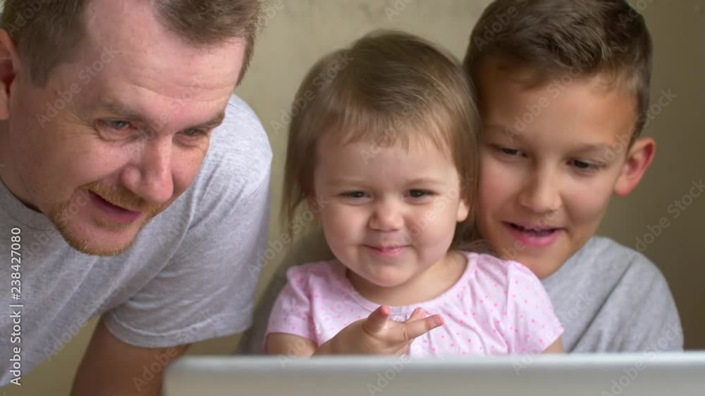 Parent and two children look at laptop screen having fun together at home. Father, son and daughter watch animated film laughing. Man and siblings, big brother, small sister smile looking to display Parent and two children look at laptop screen having fun together at home. Father, son and daughter watch animated film laughing. Man and siblings, big brother, small sister smile looking to display