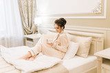 Beautiful young girl in Golden pajamas in a luxurious room with stylish hair and makeup lying in bed with a Cup of coffee. Morning bride.