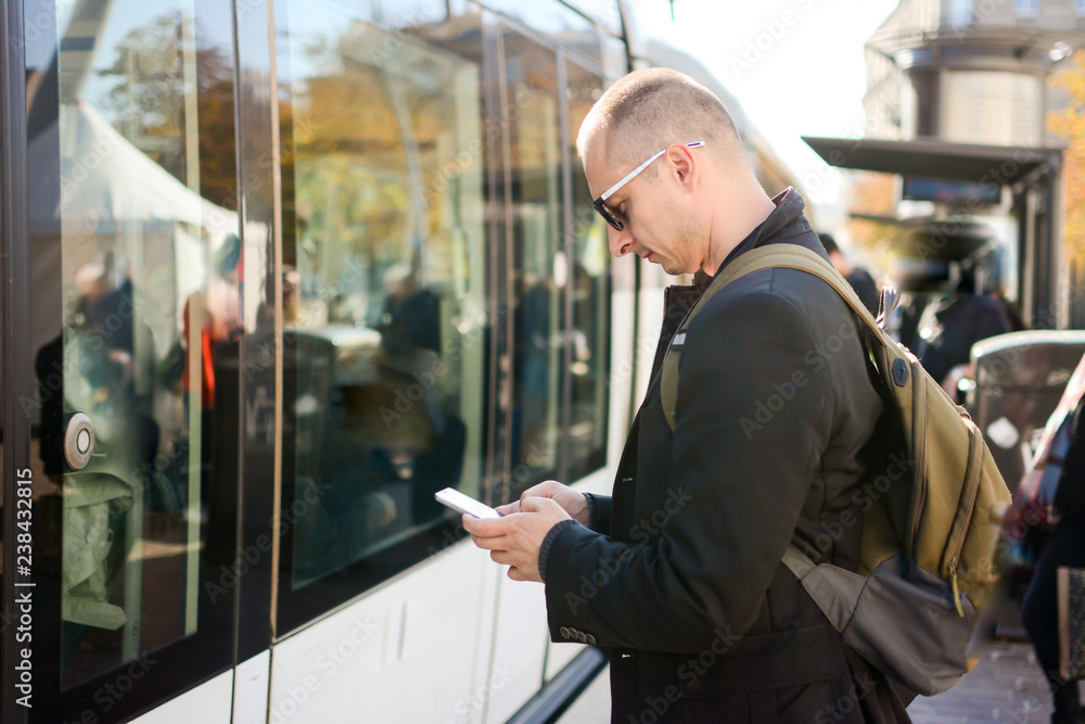 Man checking table of city traffic online on web page using smartphone, satisfied with app for paying for electric transport via cellphone waiting for tram.