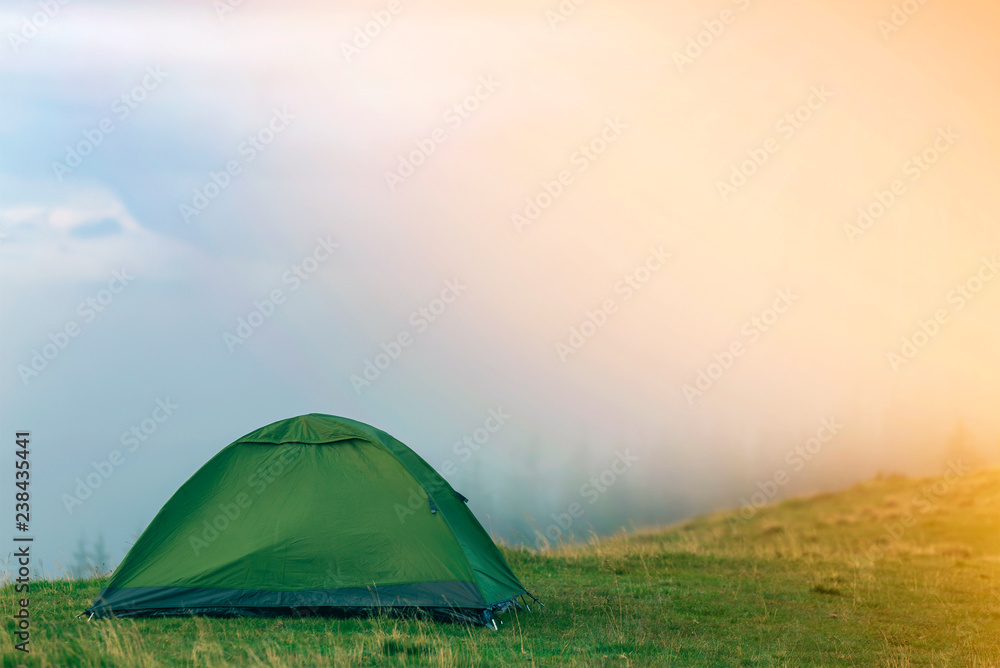 Small tourist tent on grassy mountain hill on fogy tree tops and clear blue sky before sunrise copy space background. Summer camping in mountains at dawn. Tourism, hiking and beauty of nature concept.