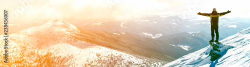 Obraz na plátne Silhouette of alone tourist standing on snowy mountain top in winner pose with raised hands enjoying view and achievement on bright sunny winter day