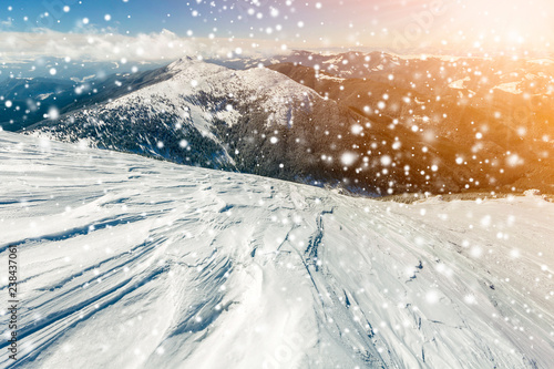 Beautiful winter landscape. Steep mountain hill slope with white deep snow, distant mountain range panorama, large snowflakes and bright shining sun rays on blue sky colorful copy space background. © bilanol