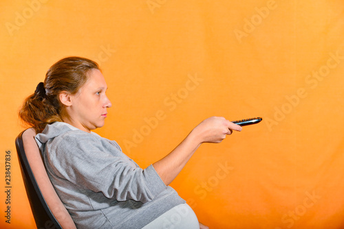 A pregnant woman sits in a chair and switches channels with a TV remote control.