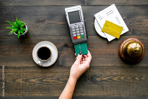 Pay the bill by payment terminal. Woman's hand insert bank card in payment terminal near bill, service bell, coffee and donuts on dark wooden background top view copy space