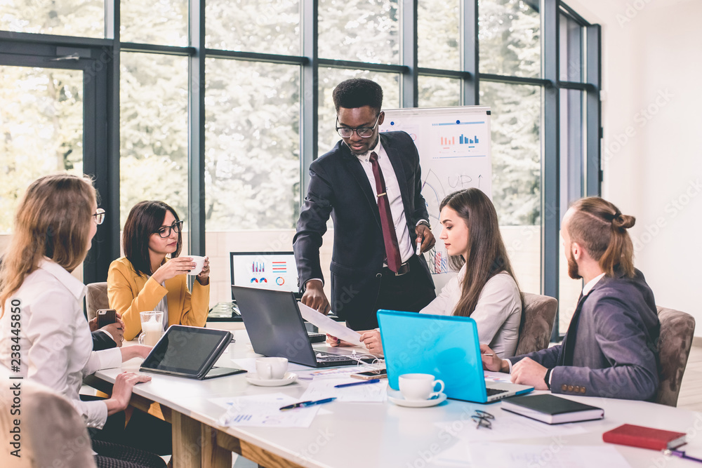 Discussing new business ideas. Cheerful young Afro American Man standing and makes a report near whiteboard while his colleagues sitting at the desk Discussing new business ideas. Cheerful young Afro American Man standing and makes a report near whiteboard while his colleagues sitting at the desk
