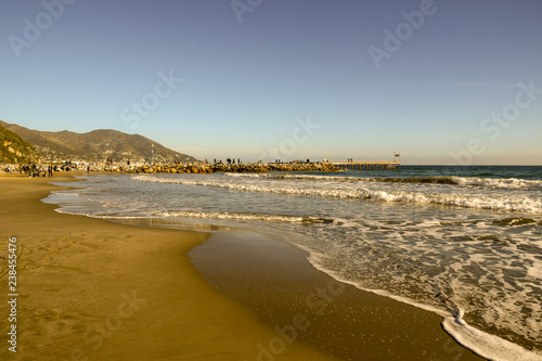 Sandy beach with people walking on a rocky dock in a sunny winter day, Laigueglia, Liguria, Italy