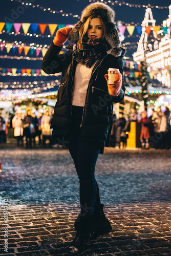 Beautiful girl with a traditional Russian hat and a glass of glintwine in her hands on the background of lights garlands