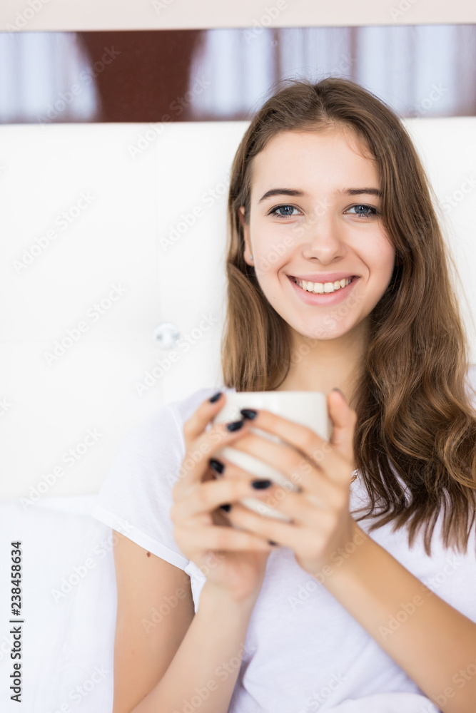 Young smiling attractive brunette woman lies in a big white bed and drinks coffee