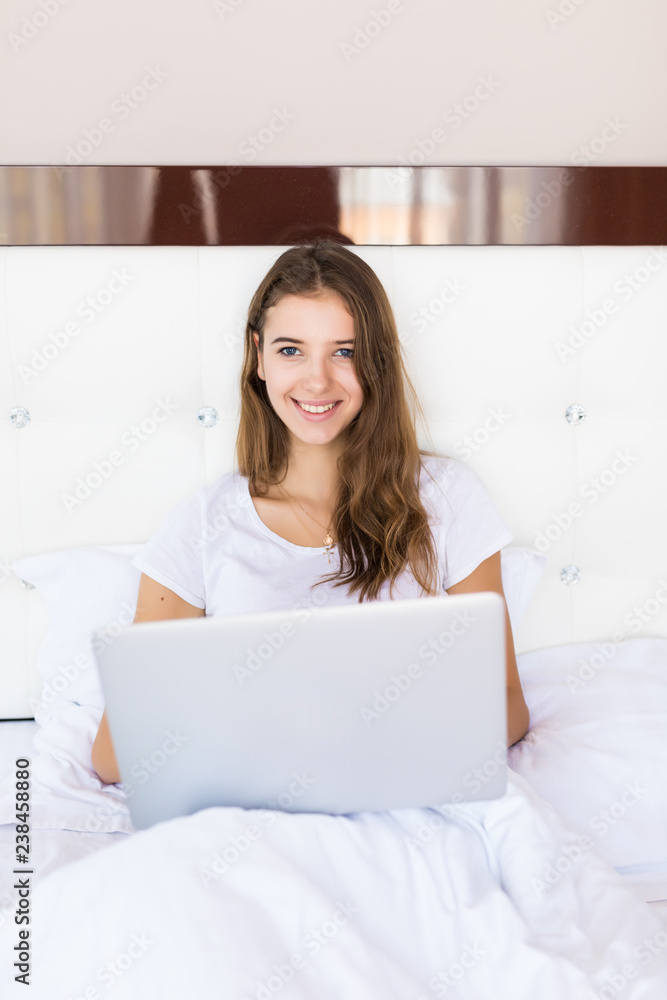Happy young brunette woman using laptop while sitting on bed in the morning