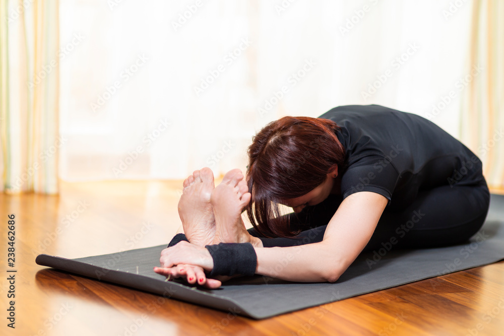 Yoga Concept. Caucasian Woman Practicing Yoga Exercise Indoors At Bright Afternoon. Sitting in Pashchimottasana Pose During Solitude Meditation Session. Equipped In Black Top Bra and Black Pants Yoga Concept. Caucasian Woman Practicing Yoga Exercise Indoors At Bright Afternoon. Sitting in Pashchimottasana Pose During Solitude Meditation Session. Equipped In Black Top Bra and Black Pants