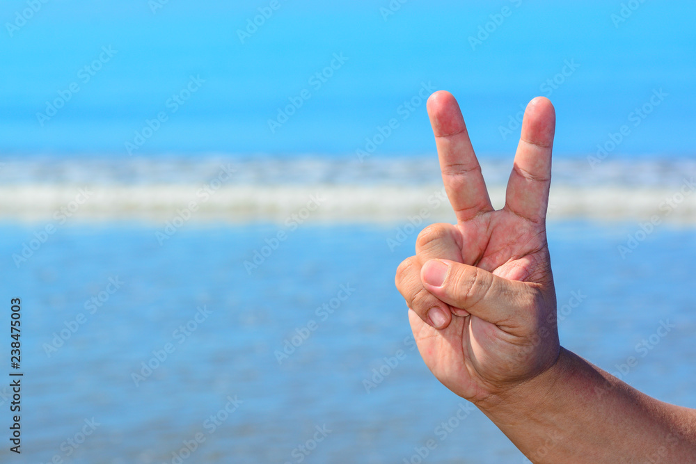 A male hand shows a peace symbol sign with two fingers isolated against blue sky and beach background. A male hand shows a peace symbol sign with two fingers isolated against blue sky and beach background.