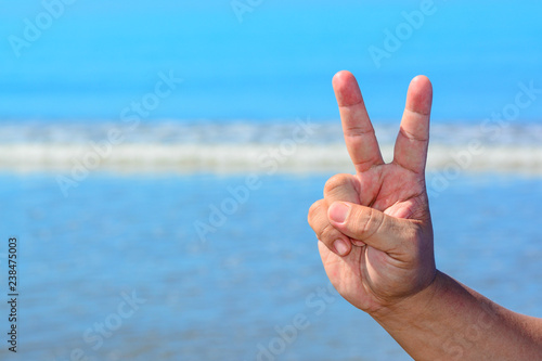 A male hand shows a peace symbol sign with two fingers isolated against blue sky and beach background. © SURIYATI