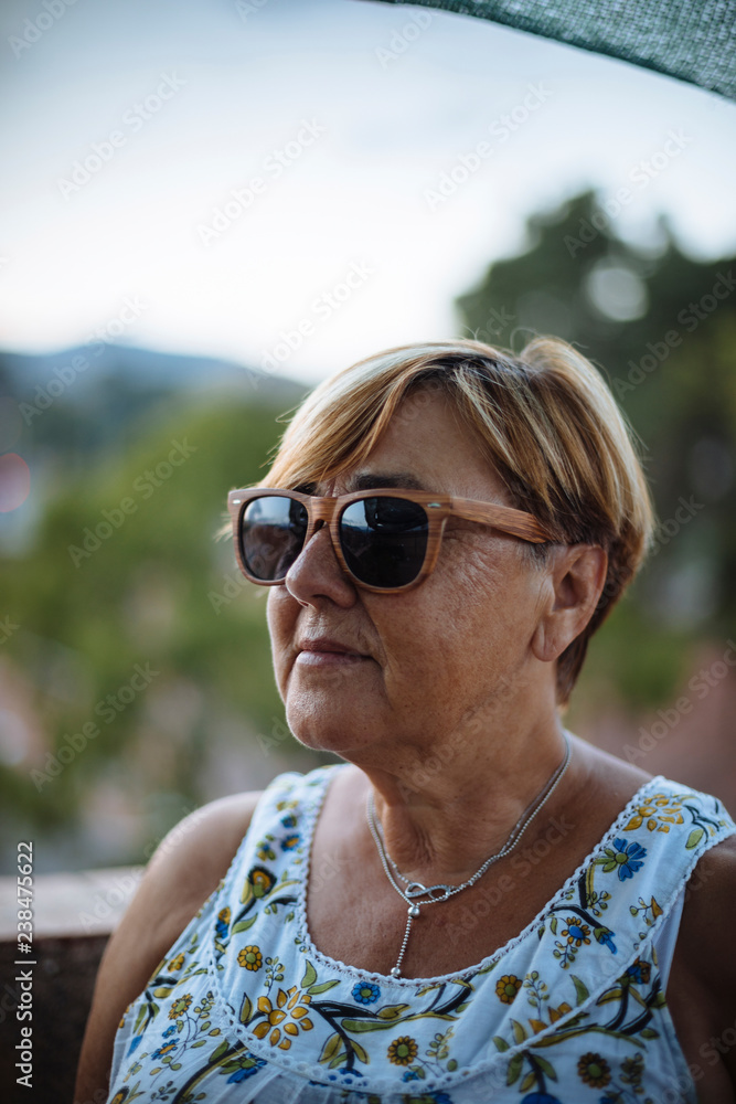 Close-up of senior woman wearing sunglasses while standing on building terrace against sky Close-up of senior woman wearing sunglasses while standing on building terrace against sky