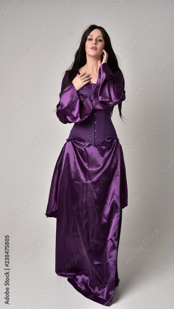 full length portrait of beautiful girl with long black hair,   wearing purple fantasy medieval gown. standing pose on grey studio background.