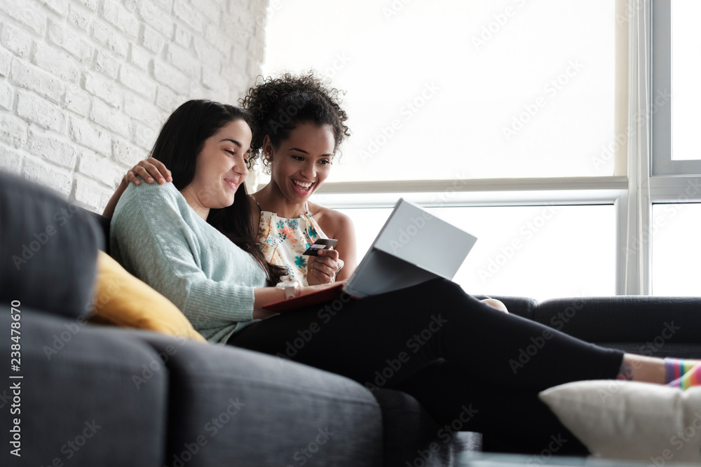 Low angle view of happy lesbian couple doing online shopping while sitting on sofa at home Low angle view of happy lesbian couple doing online shopping while sitting on sofa at home