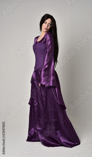 full length portrait of beautiful girl with long black hair, wearing purple fantasy medieval gown. standing pose on grey studio background.