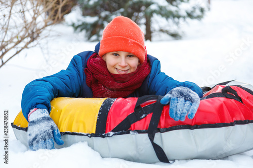cheerful cute young boy in hat red scarf and blue jacket lays on tube on snow, has fun, smiles. Teenager on sledding in winter park. Active lifestyle, winter activity, outdoor winter games, snowballs