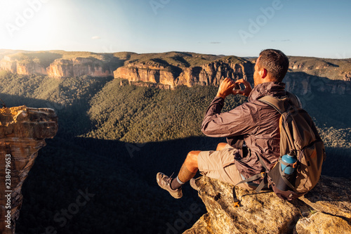 Side view of male hiker with backpack photographing while sitting on mountain against sky during sunny day photo