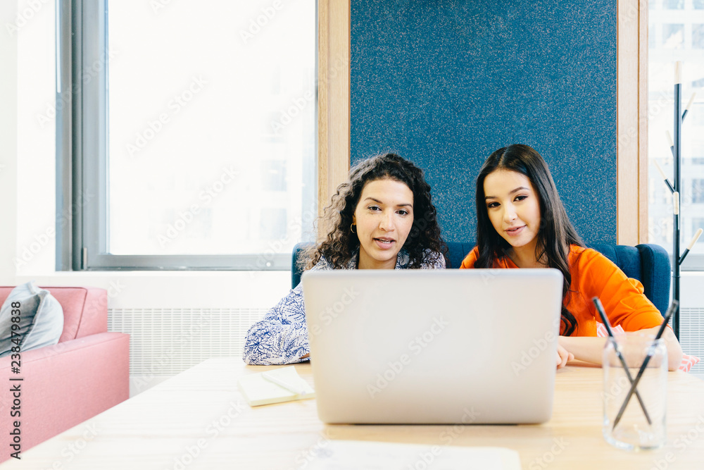 Female colleagues discussing over laptop computer while sitting at desk in office Female colleagues discussing over laptop computer while sitting at desk in office